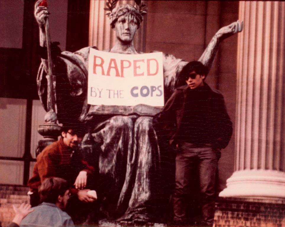 Alma Mater statue with two young men posing in front of her with a sign on her lap saying "Raped By The Cops."