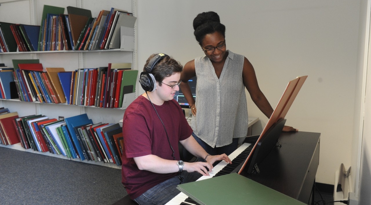 2 students in the Digital Music Lab - 1 is playing a digital piano with headphones on while the other looks on