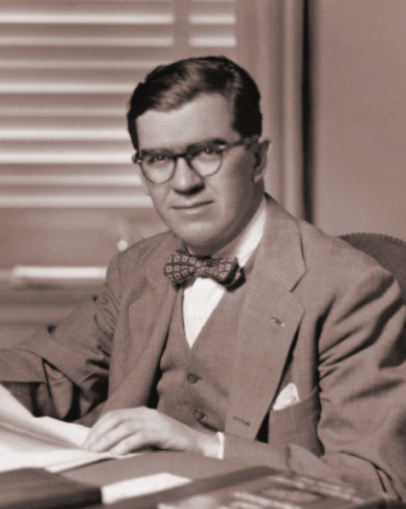 black and white portrait photo of Charles Dollard sitting at a desk in a suit holding papers